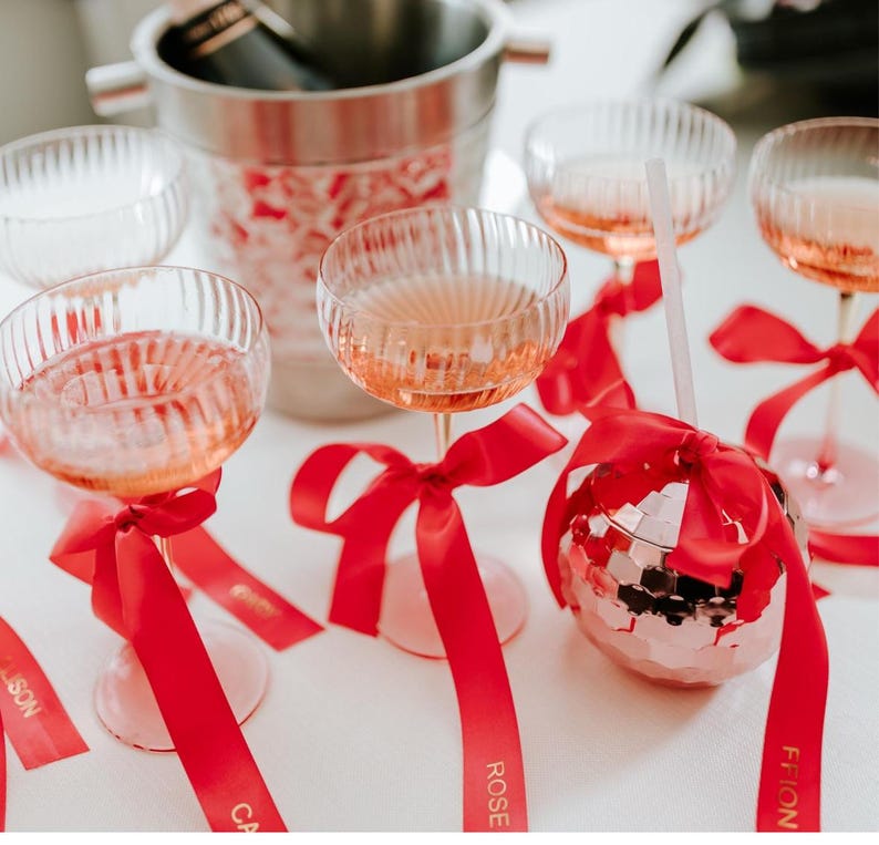 May include: A festive arrangement featuring champagne glasses filled with a pink beverage, adorned with red ribbons. A disco ball-shaped glass and a champagne bucket are also present, creating a celebratory atmosphere. The glasses have a ribbed design.