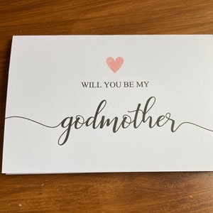 May include: A white card with a pink heart and the text "Will you be my godmother" in black cursive writing.