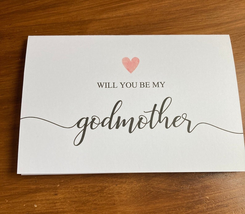 May include: A white card with a pink heart and the text "Will you be my godmother" in black cursive writing.