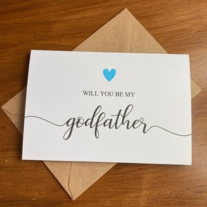 May include: A white card with a blue heart and the text "Will you be my godfather" in black cursive writing.