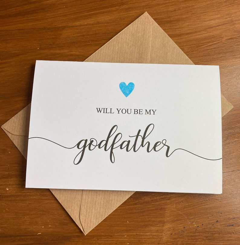 May include: A white card with a blue heart and the text "Will you be my godfather" in black cursive writing.