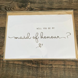May include: White card with the text "Will you be my maid of honour?" in black cursive writing with a small heart drawn below the text.
