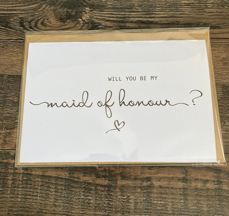 May include: White card with the text "Will you be my maid of honour?" in black cursive writing with a small heart drawn below the text.