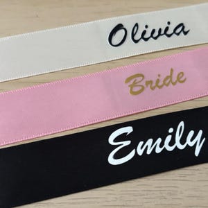May include: Close-up of three ribbons with text. The top ribbon is off-white with the name "Olivia" in black script. The middle ribbon is pink with the word "Bride" in gold. The bottom ribbon is black with the name "Emily" in white.
