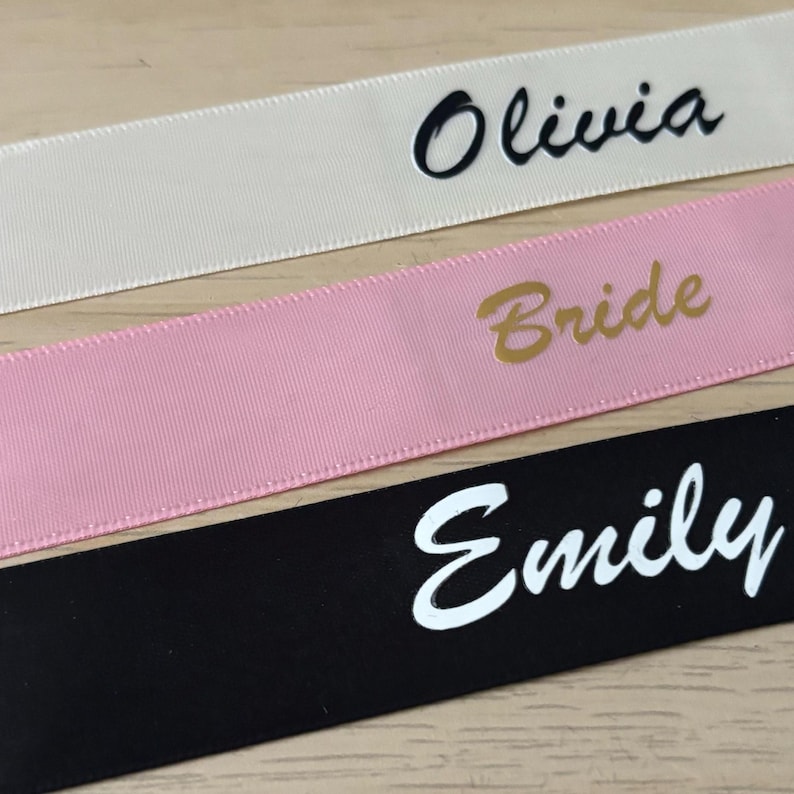 May include: Close-up of three ribbons with text. The top ribbon is off-white with the name "Olivia" in black script. The middle ribbon is pink with the word "Bride" in gold. The bottom ribbon is black with the name "Emily" in white.