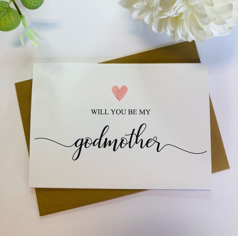 May include: A white card with a pink heart and the text "Will you be my godmother" in black cursive writing.