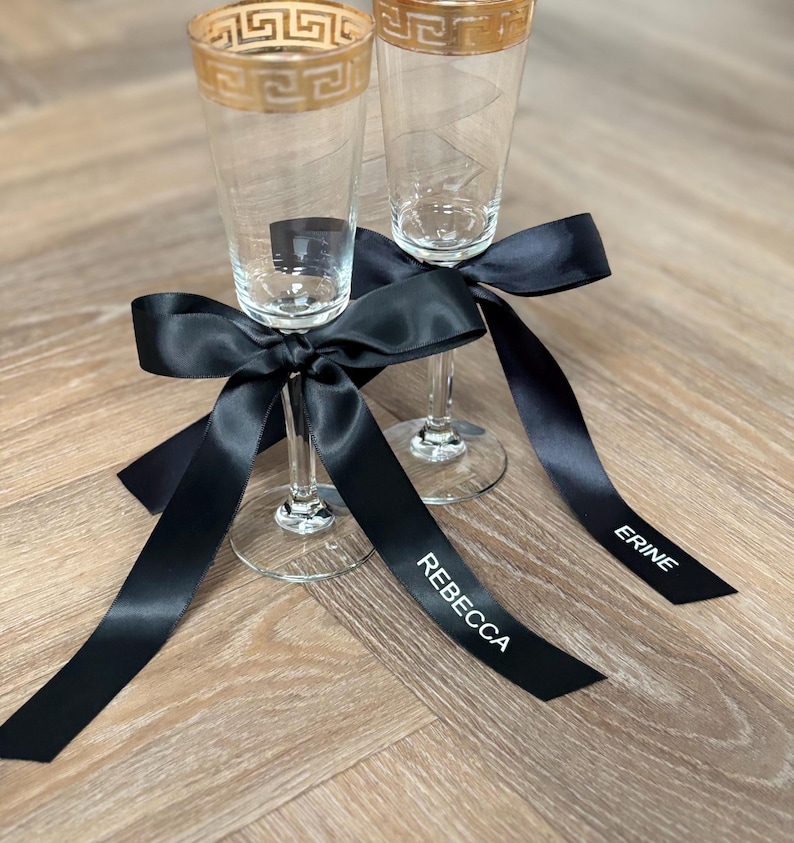 May include: Two champagne flutes with gold rims and a Greek key design are adorned with black satin ribbons. The ribbons are tied in bows and feature the names "Rebecca" and "Erine" in white lettering, perfect for a wedding or special event.