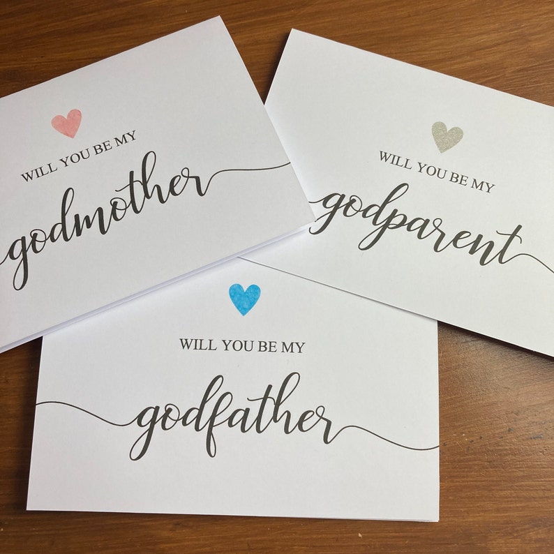 May include: Three white cards with the text "Will you be my..." followed by "godmother", "godparent", and "godfather". Each card has a heart in a different color: pink, gray, and blue.