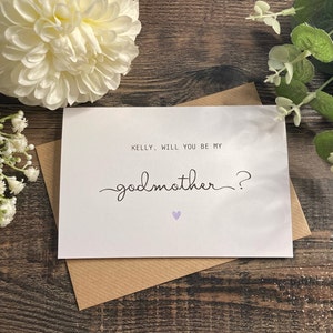 Pu&ograve; includere: Un biglietto bianco con una busta marrone. Il biglietto ha il testo "KELLY, WILL YOU BE MY GODMOTHER?" con un piccolo cuore viola.