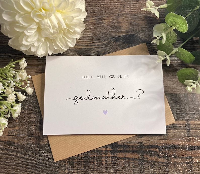 Pu&ograve; includere: Un biglietto bianco con una busta marrone. Il biglietto ha il testo "KELLY, WILL YOU BE MY GODMOTHER?" con un piccolo cuore viola.