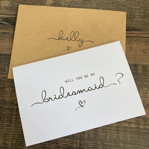 May include: A white card with the text "Will you be my bridesmaid?" in black cursive writing. A small heart is drawn below the text. The card is on top of a brown envelope with the name "Kelly" written in black cursive writing on the front.