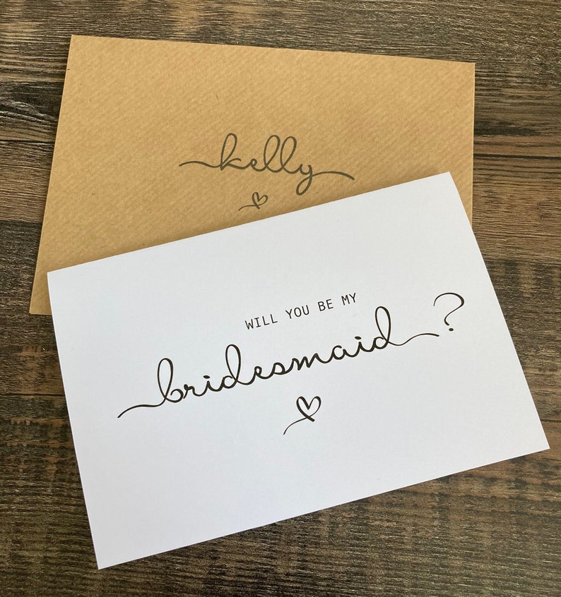 May include: A white card with the text "Will you be my bridesmaid?" in black cursive writing. A small heart is drawn below the text. The card is on top of a brown envelope with the name "Kelly" written in black cursive writing on the front.