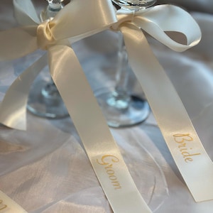 May include: Two champagne flutes adorned with ivory satin ribbons. The ribbons are tied into bows and feature the words "Groom" and "Bride" in gold lettering, perfect for a wedding celebration.