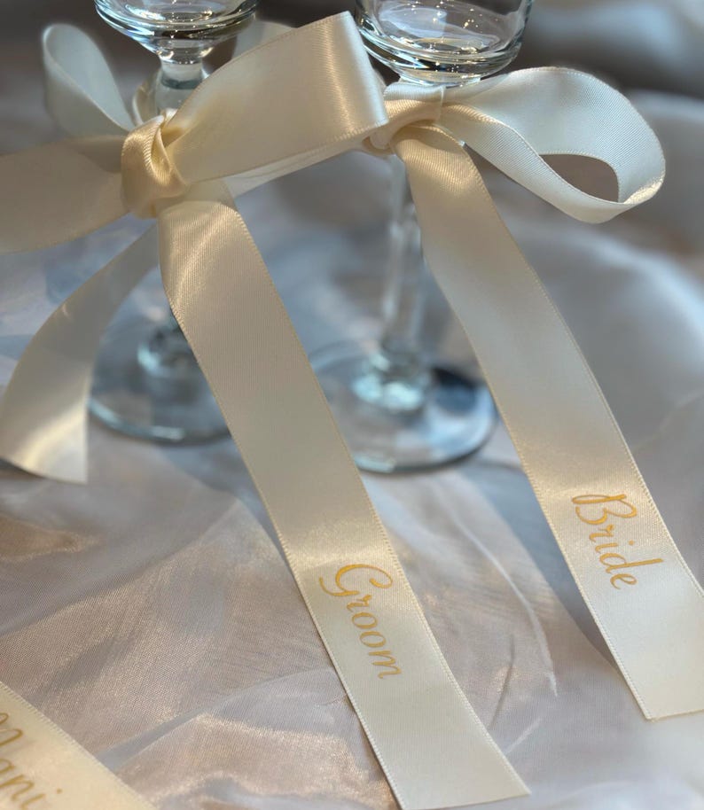 May include: Two champagne flutes adorned with ivory satin ribbons. The ribbons are tied into bows and feature the words "Groom" and "Bride" in gold lettering, perfect for a wedding celebration.