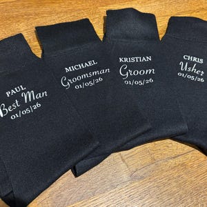 May include: Four pairs of black socks with white text. Each pair has a different title: "Best Man", "Groomsman", "Groom", and "Usher", along with a date. The socks are arranged on a wooden surface.