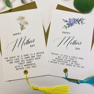 May include: Two white cards with gold trim and tassels. The cards feature a floral design and the text "Happy Mother's Day". The first card has a quote about the heart of a home being a mother. The second card has a quote about the meaning of the word "mother".