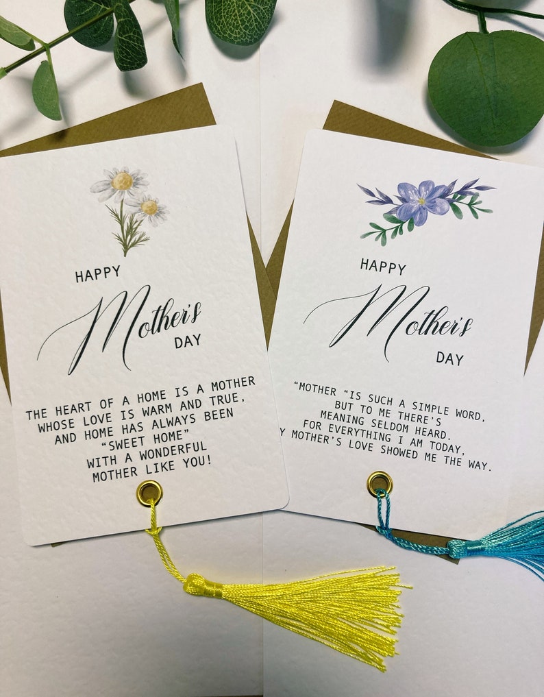 May include: Two white cards with gold trim and tassels. The cards feature a floral design and the text "Happy Mother's Day". The first card has a quote about the heart of a home being a mother. The second card has a quote about the meaning of the word "mother".