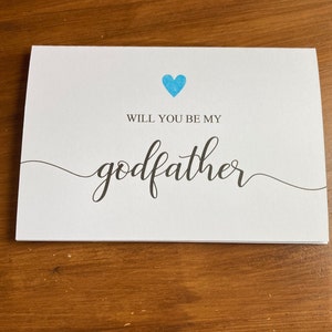May include: A white card with a blue heart and the text "Will you be my godfather?" in black cursive font.