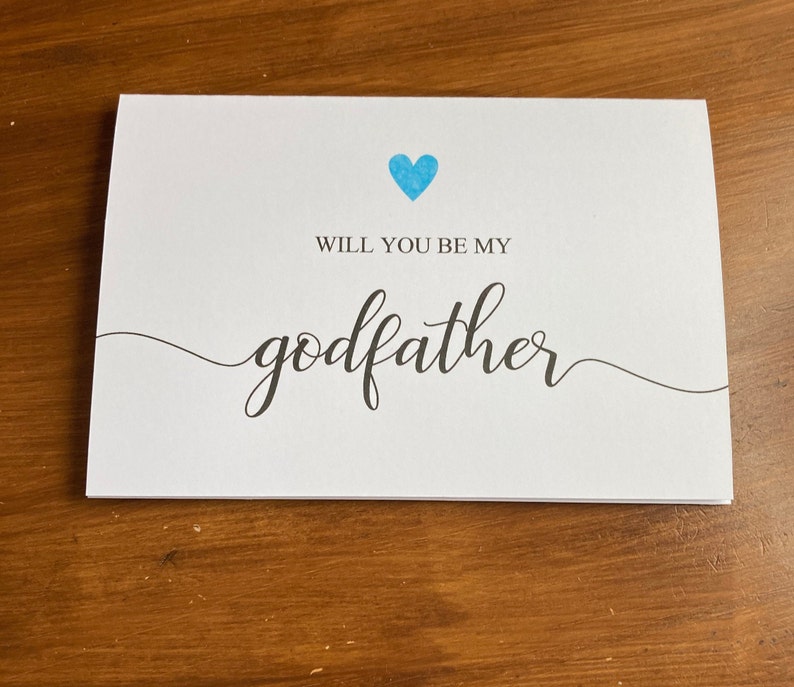 May include: A white card with a blue heart and the text "Will you be my godfather?" in black cursive font.