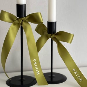 May include: Two black metal candle holders with white candles. Each candle holder has a green ribbon tied in a bow. One ribbon says "GROOM" and the other says "BRIDE" in white text. Wedding decor.