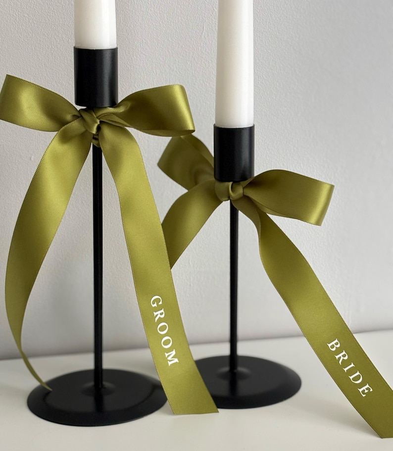 May include: Two black metal candle holders with white candles. Each candle holder has a green ribbon tied in a bow. One ribbon says "GROOM" and the other says "BRIDE" in white text. Wedding decor.