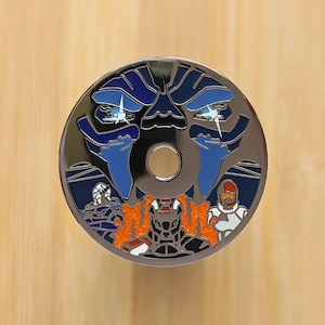 May include: A circular, silver-toned pin with a robot-themed design. The artwork features a central hole and a detailed illustration with blue and orange colours. The design includes a stylized face.