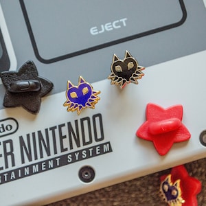 May include: A close-up of a Super Nintendo Entertainment System with four pins on top. The pins are shaped like stars and feature a black cat, a blue cat, and two red stars. The text "SUPER NINTENDO ENTERTAINMENT SYSTEM" is visible on the console.
