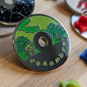 May include: A round, silver-coloured pin with a green and black design. The top half of the pin features a green landscape with trees and clouds. The bottom half of the pin is black and features a series of small, silver-coloured characters.