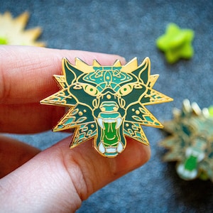 May include: A green and gold enamel pin featuring a stylized wolf head with a detailed design. The wolf's mouth is open and its fangs are visible. The pin is a popular collectible for fans of fantasy and mythology.