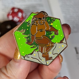May include: A green and gold enamel pin featuring a brown figure with a leafy crown standing on a green and white background.