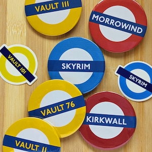 May include: Nine round, colorful, pinback buttons with various video game and fantasy themed designs. The buttons feature the text "Vault III", "Morrowind", "Skyrim", "Vault 101", "Vault 76", "Kirkwall", and "Skyrim" in a circular design.