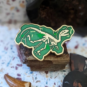 May include: A gold-coloured enamel pin featuring a white dinosaur skeleton on a green background. The dinosaur is in a resting position with its legs and tail spread out. The pin is surrounded by a green border.