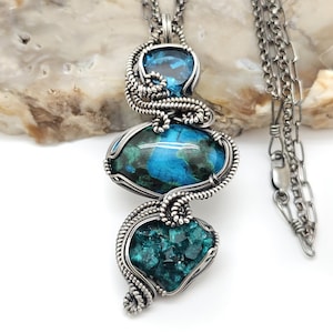 May include: A handcrafted silver pendant necklace featuring three gemstone elements. The top stone is a clear blue, the middle stone is a vibrant blue and green, and the bottom stone is a deep teal. The stones are encased in a detailed silver wire design.