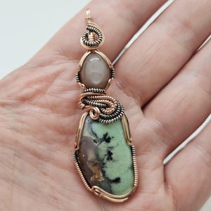 May include: A handcrafted pendant featuring a light green and brown stone, accented with black inclusions. The pendant is wire-wrapped with copper and silver-toned metal, and includes a gray oval stone. The design is intricate and detailed.