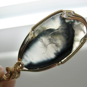 May include: A gold wire wrapped pendant featuring a gray and black agate stone with a unique pattern. The stone is oval shaped and has a smooth, polished finish.