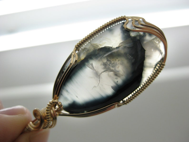 May include: A gold wire wrapped pendant featuring a gray and black agate stone with a unique pattern. The stone is oval shaped and has a smooth, polished finish.