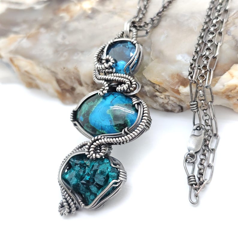 May include: A silver pendant necklace featuring three gemstone pendants in shades of blue and teal. The stones are encased in intricate wirework and suspended from a delicate chain. The necklace is displayed against a white background.