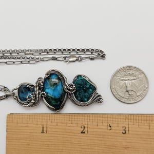 May include: A silver-toned necklace with three gemstone pendants. The pendants feature blue and green stones, intricately wire-wrapped. A ruler and a quarter are next to the necklace for scale. The necklace is on a white background.