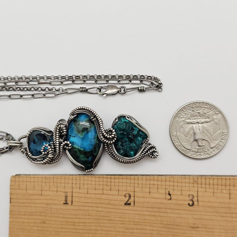 May include: A silver-toned necklace with three gemstone pendants. The pendants feature blue and green stones, intricately wire-wrapped. A ruler and a quarter are next to the necklace for scale. The necklace is on a white background.
