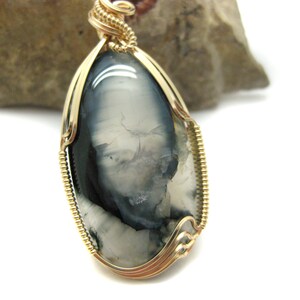 May include: A gold wire wrapped pendant featuring a gray and white agate stone with a unique pattern. The stone is oval shaped and has a smooth, polished finish.