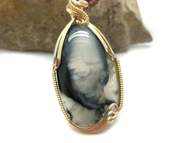 May include: A gold wire wrapped pendant featuring a gray and white agate stone with a unique pattern. The stone is oval shaped and has a smooth, polished finish.