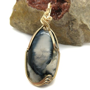 May include: A gold wire wrapped pendant featuring a gray and white agate stone. The stone has a unique pattern of swirls and veins.