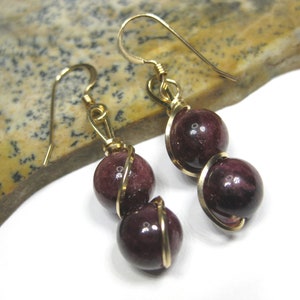 May include: A pair of gold earrings with two dark red round gemstone beads on each earring. The beads are wrapped in gold wire.
