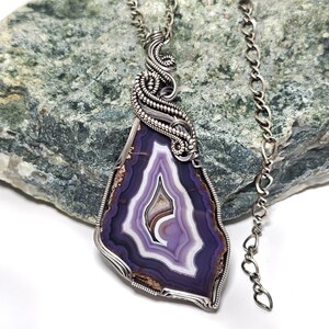 May include: A handcrafted pendant necklace featuring a polished, purple and white agate stone. The stone is encased in a silver wire wrap with intricate detailing, suspended from a silver chain. The agate displays concentric bands of color.