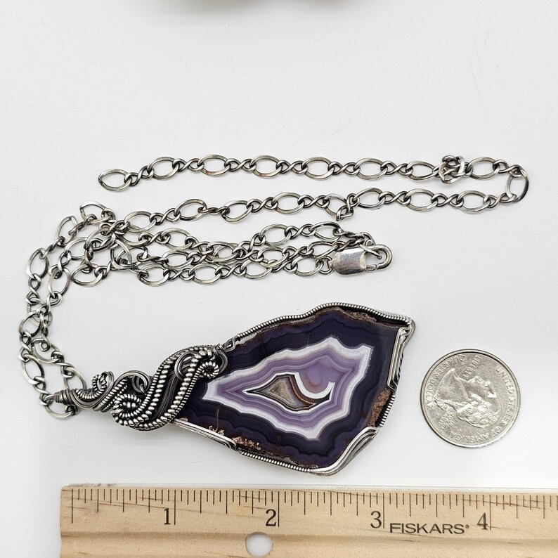 May include: A silver chain necklace with a large, polished agate pendant. The pendant features concentric bands of purple, white, and brown. A quarter coin is next to the pendant for size comparison.