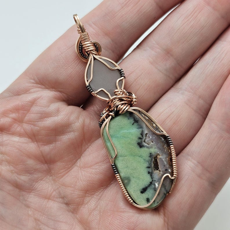 May include: A handcrafted pendant featuring two natural stones. The top stone is a light gray, teardrop shape. The bottom stone is a green and brown oval. Both are wrapped in copper wire and suspended from a gold-colored bail.