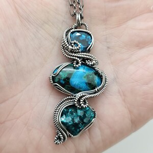 May include: A handcrafted silver pendant necklace featuring three gemstone elements. The top stone is a faceted blue gem, the middle stone is a blue and green marbled stone, and the bottom stone is a cluster of teal gems. The stones are wrapped in silver wire.