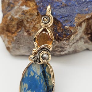 May include: A handcrafted pendant featuring a large, blue stone with a unique pattern, accented by a golden-colored metal frame and intricate wirework. The pendant also includes a faceted, amber-colored gemstone and a silver-colored bead.