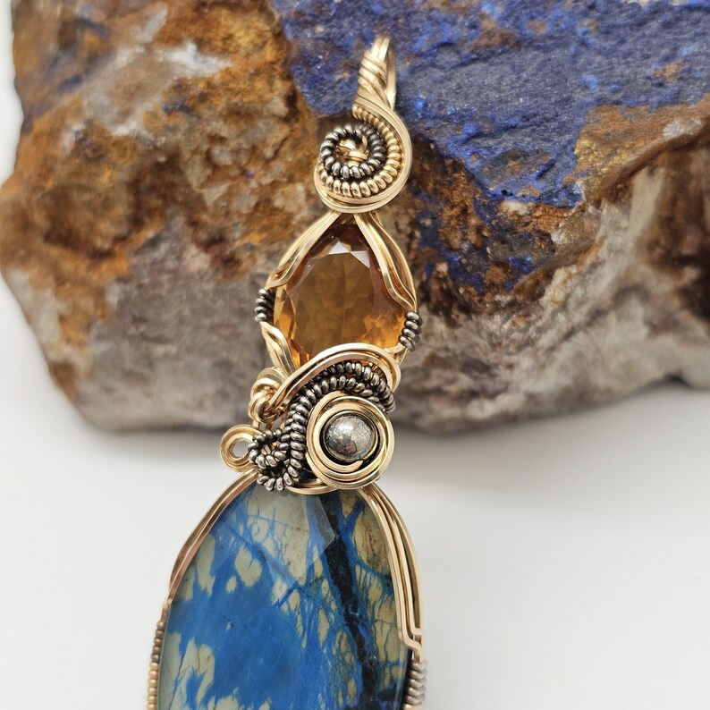 May include: A handcrafted pendant featuring a large, blue stone with a unique pattern, accented by a golden-colored metal frame and intricate wirework. The pendant also includes a faceted, amber-colored gemstone and a silver-colored bead.