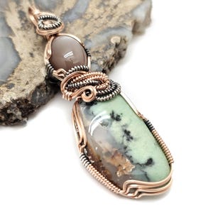 May include: A handcrafted pendant featuring a light green and brown stone, accented with black inclusions. The stone is encased in copper wire and a smaller, oval-shaped, gray stone is set above it. The pendant is displayed on a textured, gray surface.